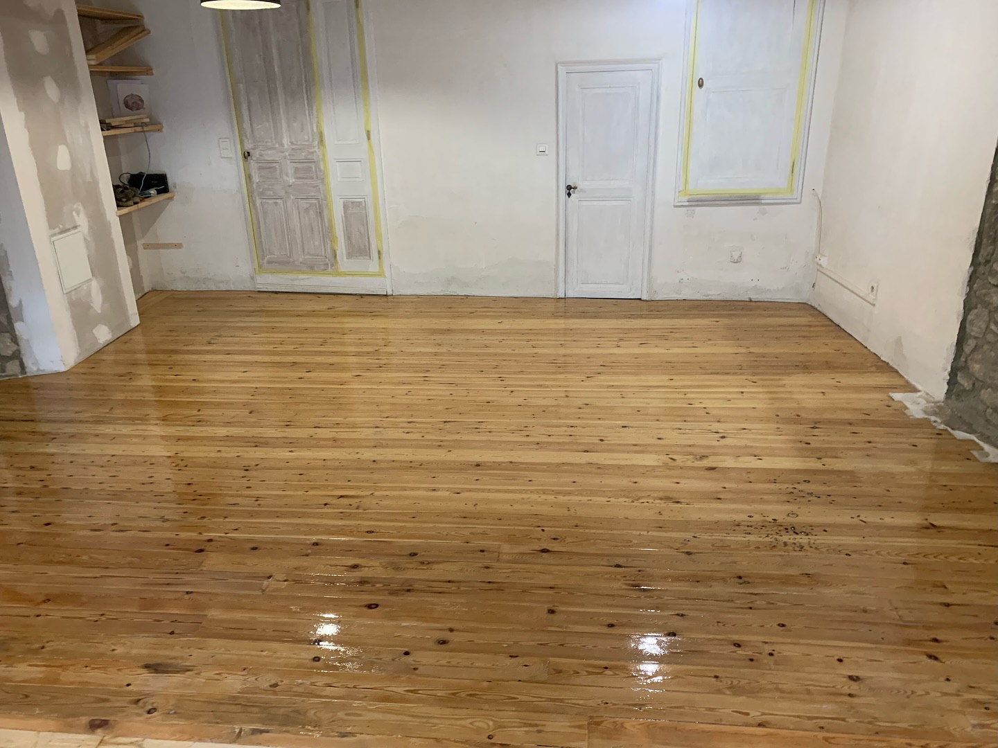 Parquet en bois de pin fraîchement verni, brillant, dans une pièce en rénovation avec portes et murs blancs bruts.