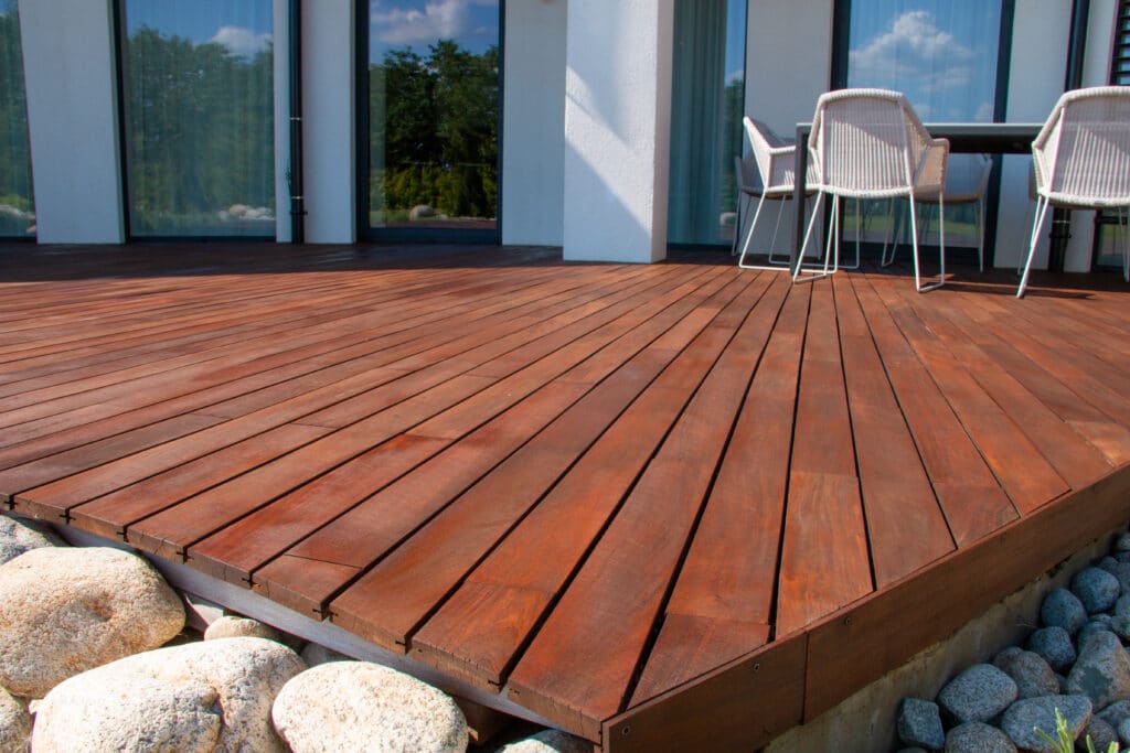 Terrasse bois rouge et mobilier extérieur moderne Terrasse en bois rouge foncé, près d'une façade blanche avec grandes baies vitrées reflétant le ciel et des arbres. Gros plan sur les galets clairs et gris.