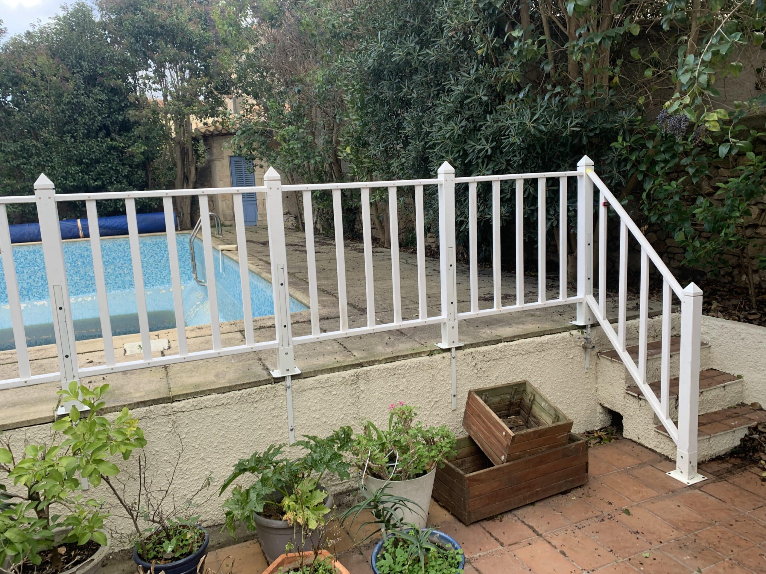 Barrière de sécurité blanche pour piscine carrelée, entourée de végétation dense et de pots de fleurs sur une terrasse en terre cuite.