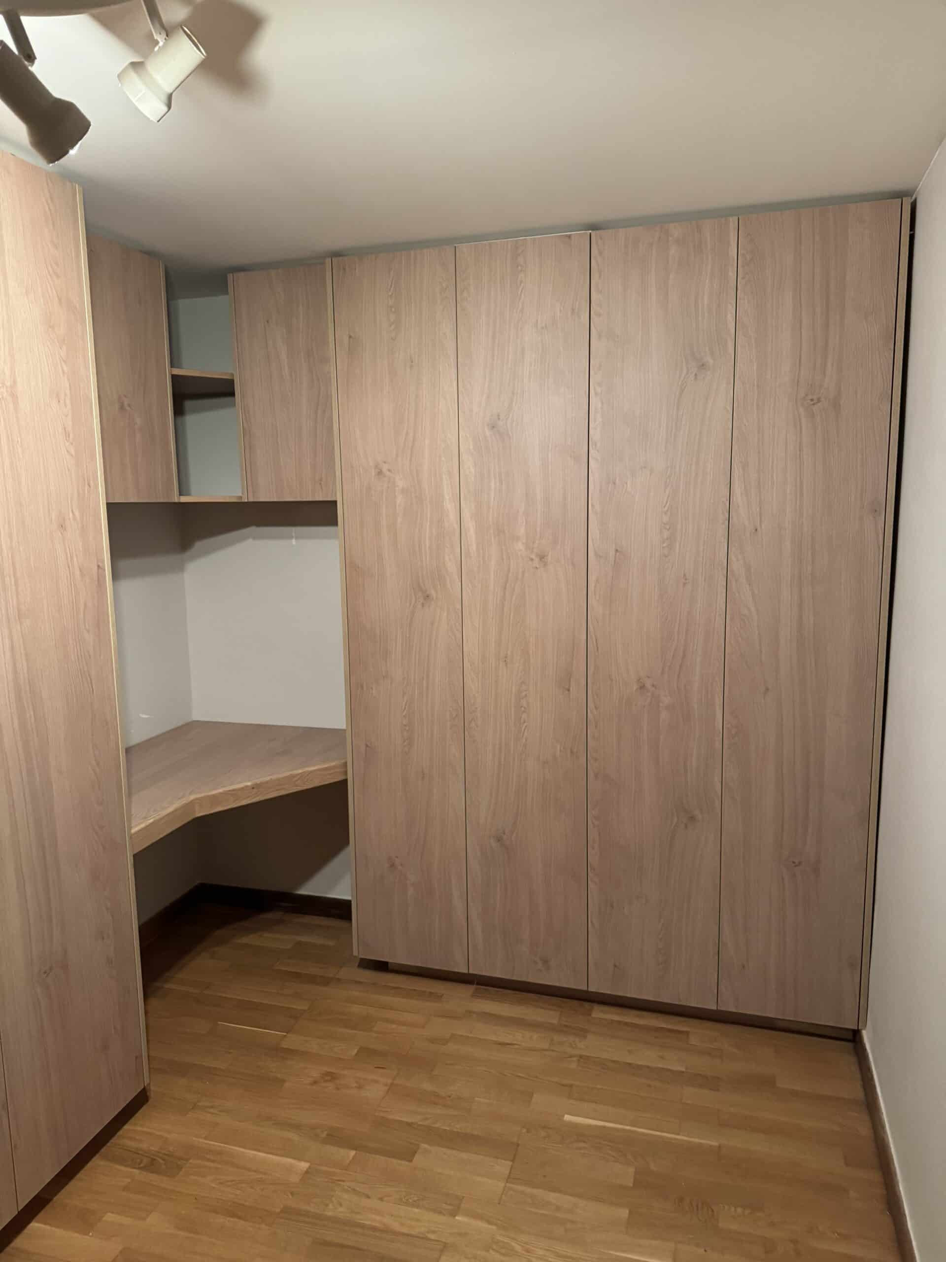 Coin dressing ou chambre avec grand placard bois clair, bureau intégré, et spots au plafond.