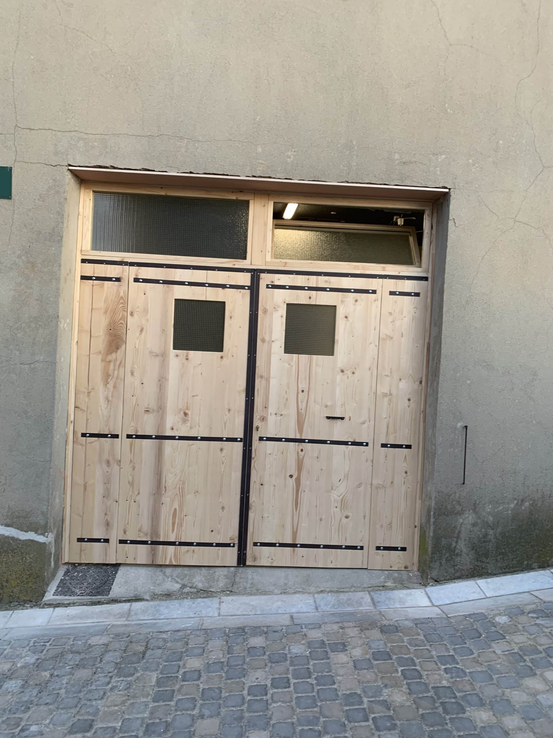 Double porte en bois clair neuve avec fenêtres grillagées, encadrée par un mur gris fissuré et un trottoir en pavés.