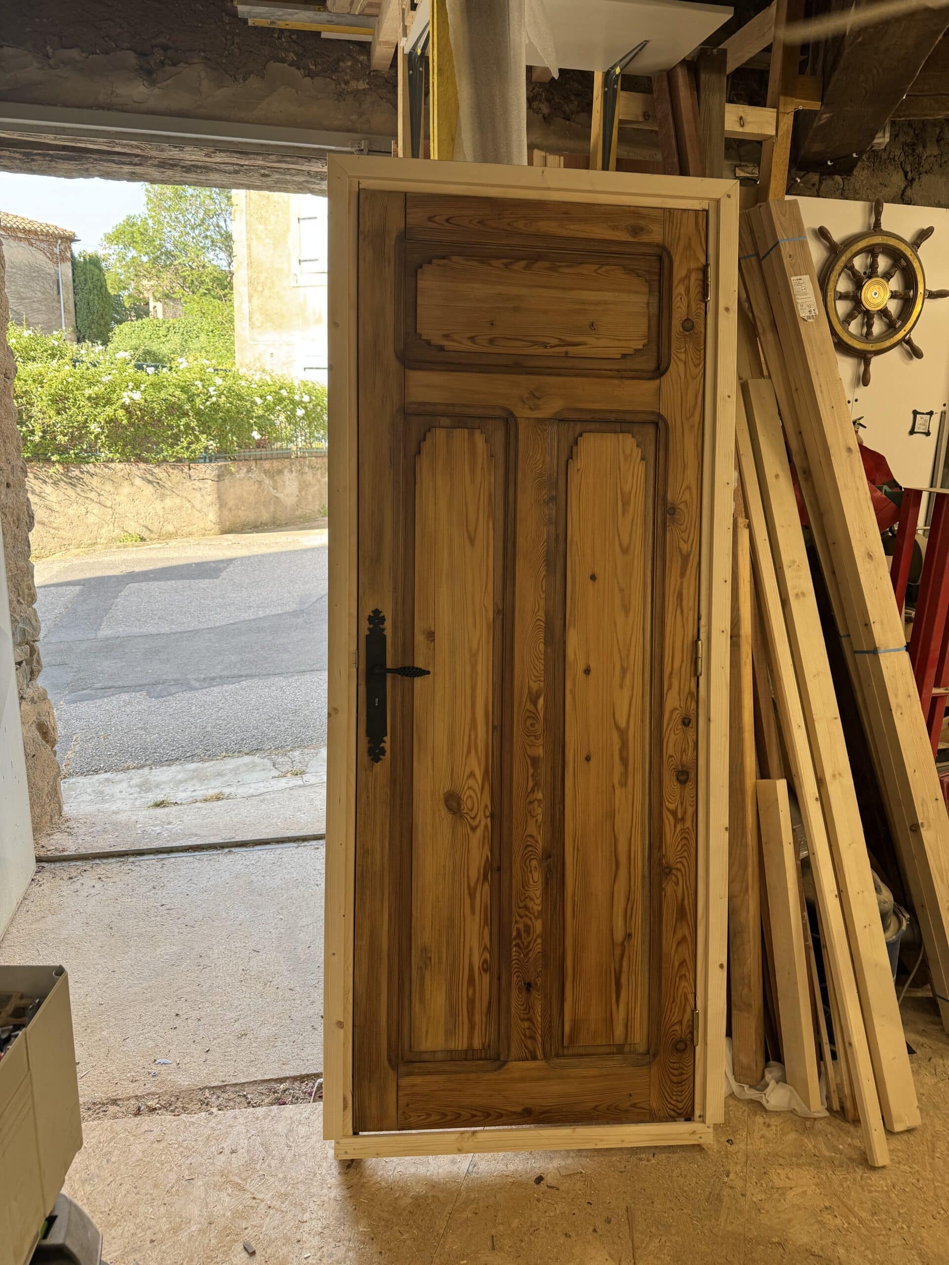 Porte ancienne en pin avec quincaillerie noire installée dans un cadre provisoire, vue depuis un intérieur de garage donnant sur une rue ensoleillée.