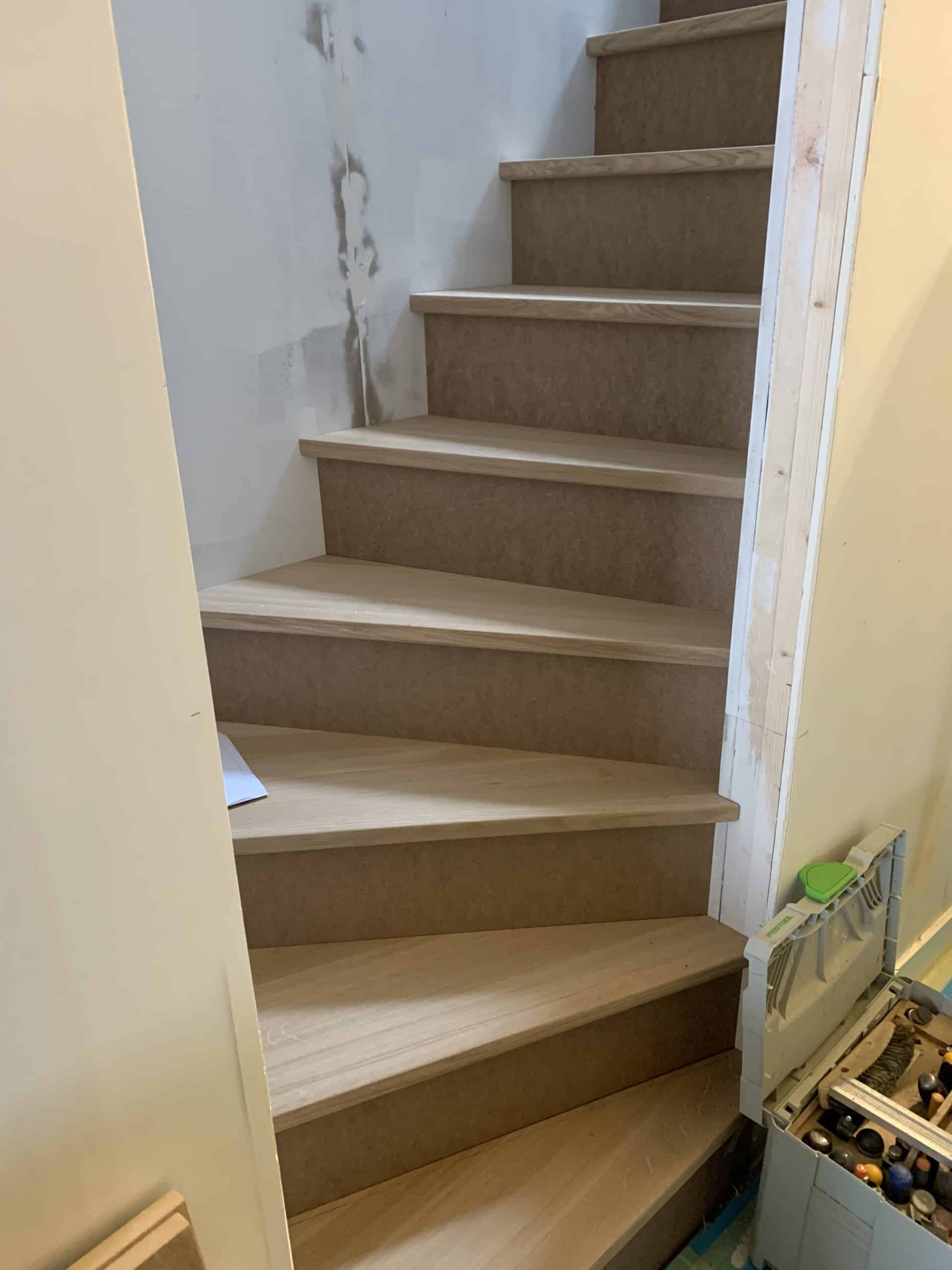 Escalier en bois clair en cours d'installation, avec contremarches en panneau de particules, près d'un mur en plâtre brut et d'une caisse à outils Festool ouverte.
