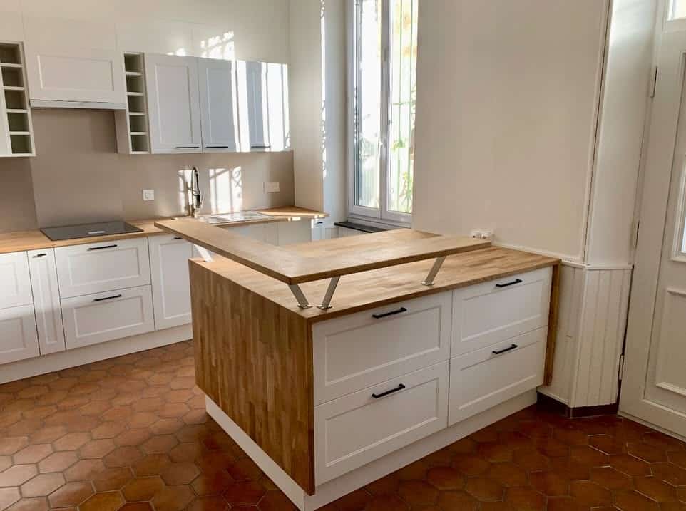 Cuisine blanche et bois avec îlot central, bar surélevé et sol en tomettes hexagonales.