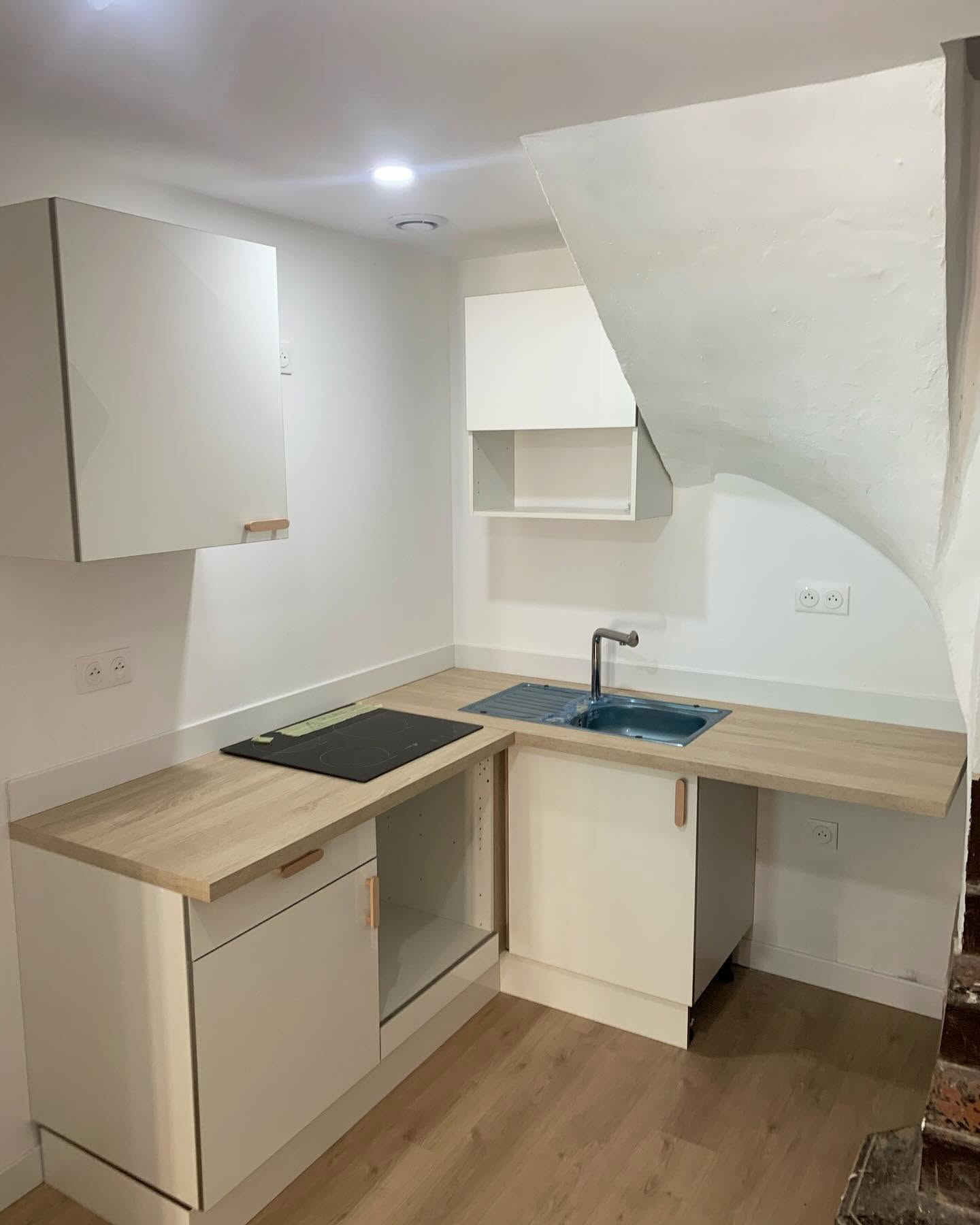Petite kitchenette moderne sous un escalier avec meubles blancs, plan de travail bois et plaque de cuisson induction.
