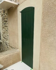Volet battant vert foncé cintré sur façade en crépi beige et mur en pierres apparentes.